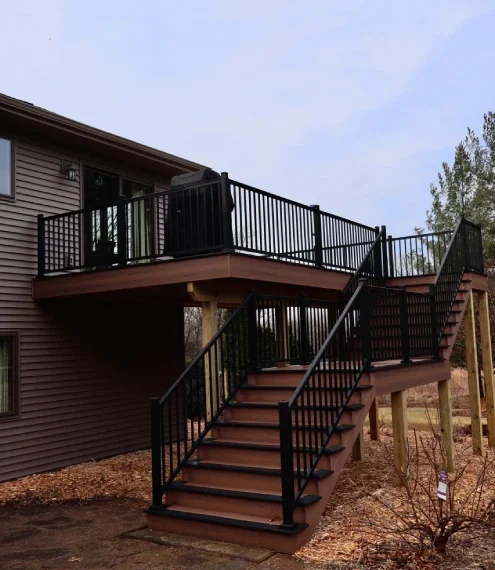 A wooden deck with a railing and stairs leading up to a house, surrounded by greenery.